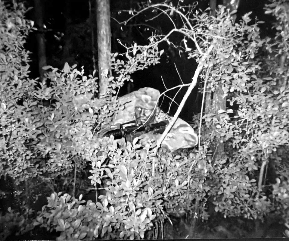 Craig Smith in camouflage with a rifle, concealed in dense brush during training at JRTC
