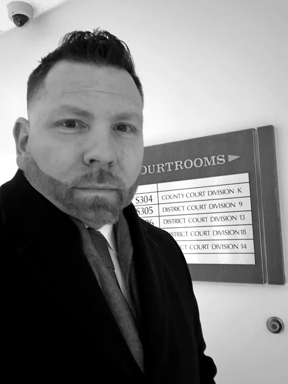 Craig Smith in a dark overcoat and tie beside a courtrooms directory sign