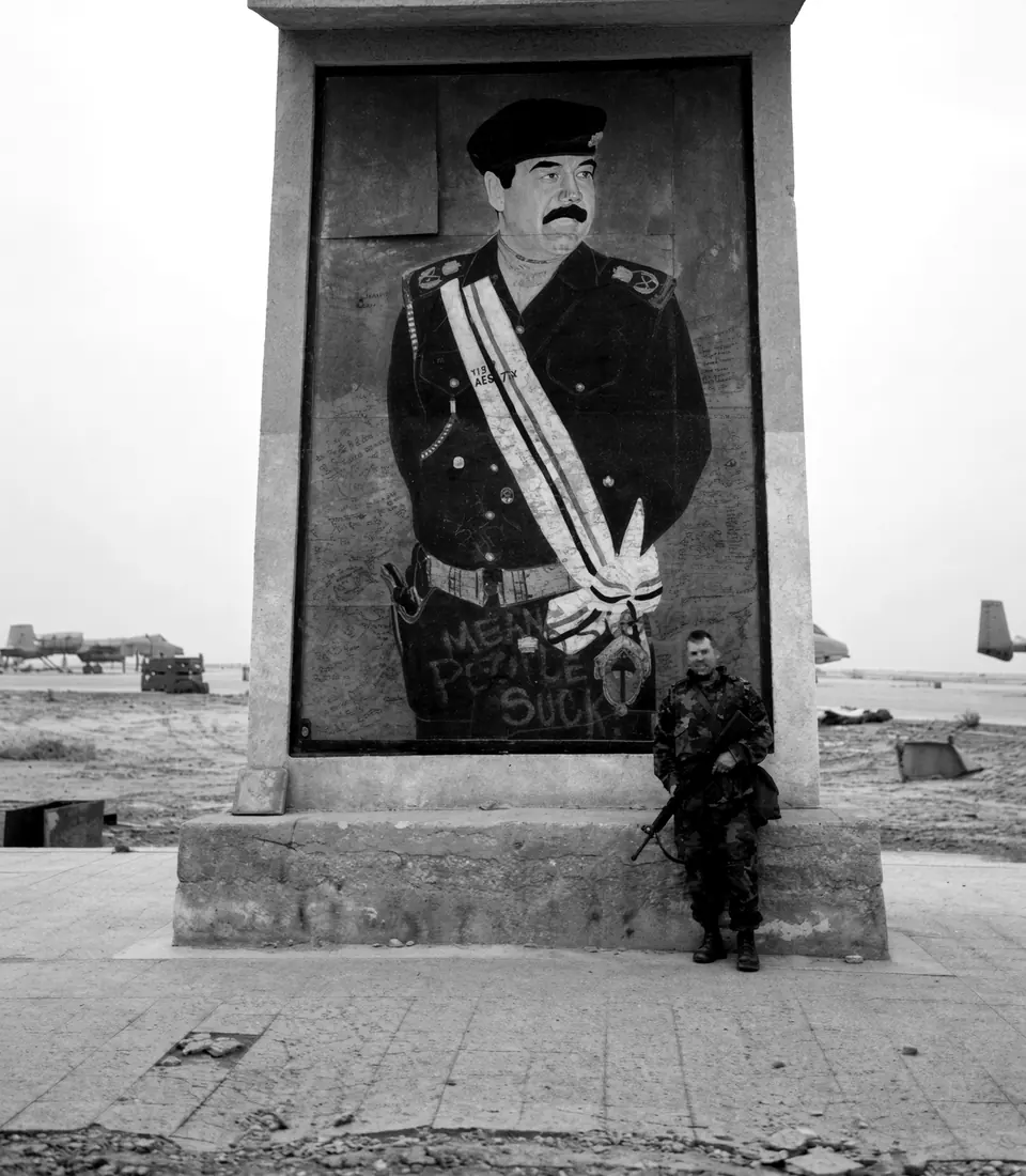 Craig Smith standing in front of a large Saddam Hussein mural at a captured airfield, aircraft in the distance