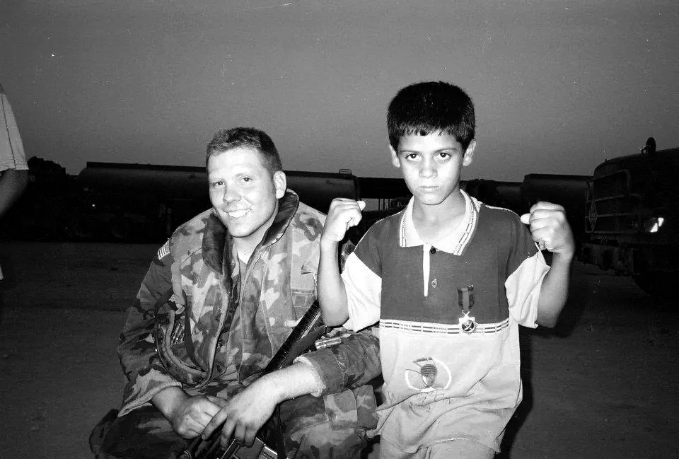 Craig Smith in uniform kneeling beside a young Iraqi boy showing his fists, military trucks behind them, Basra, Iraq